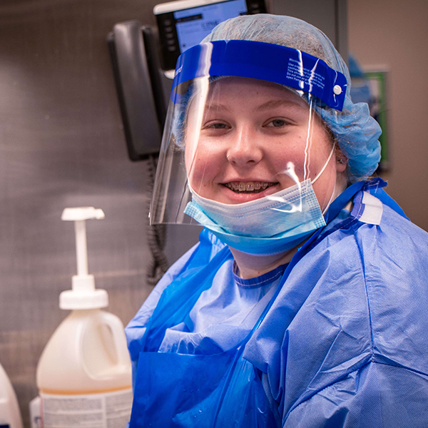 Young woman with face shield smiling inside of a clinical setting.