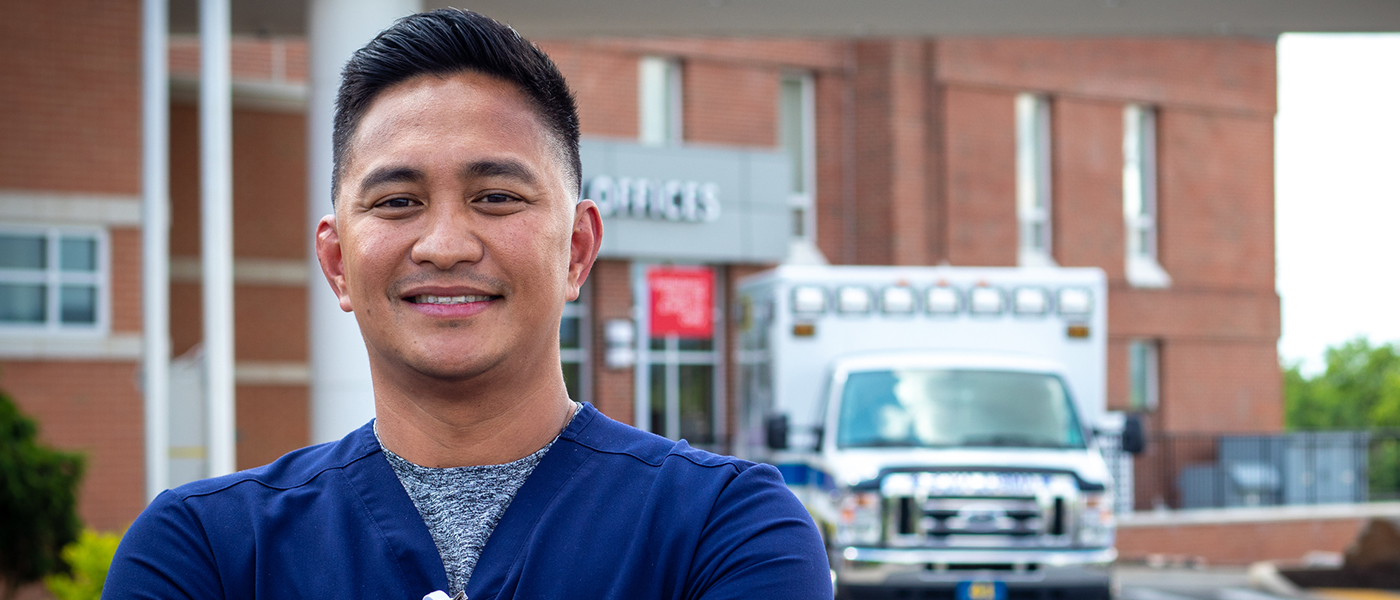 Male nurse stands in front of hospital, smiling at the camera.