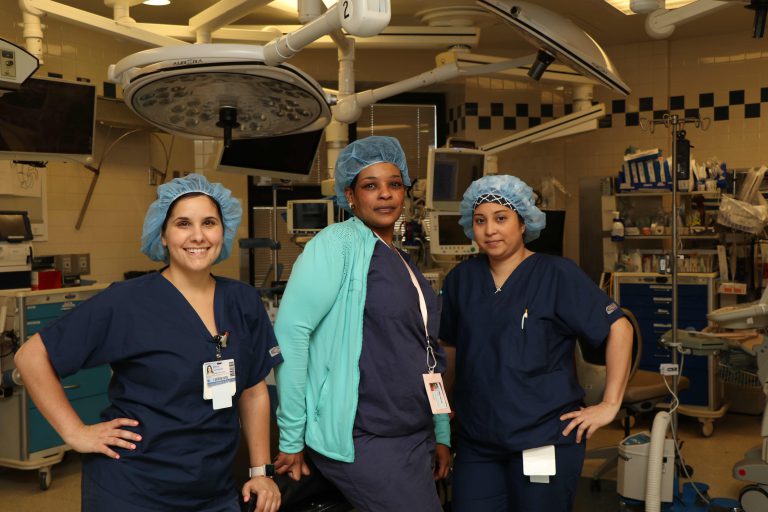 Three woman stand in operating room in scrubs.