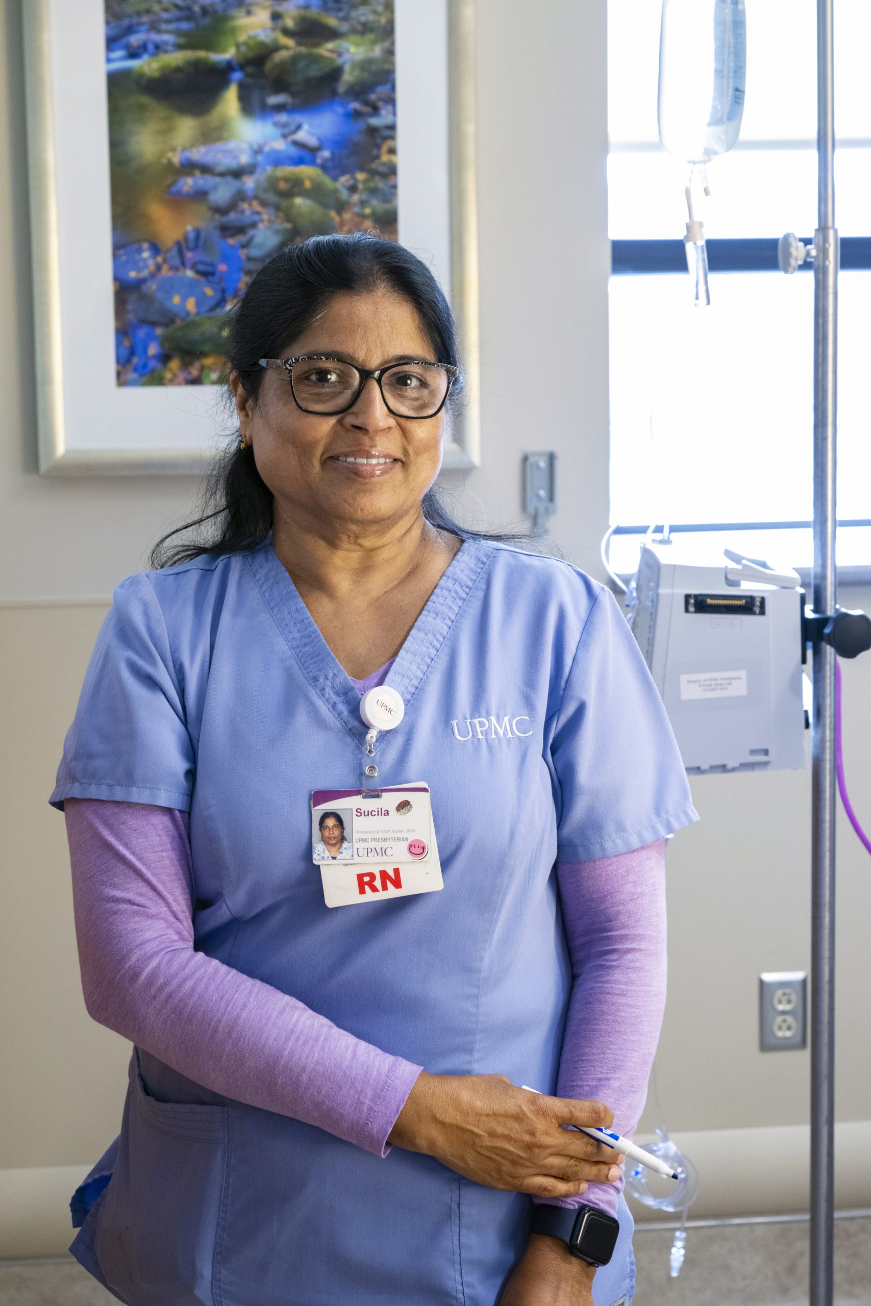 Middle aged nurse with glasses stands next to hospital bed.