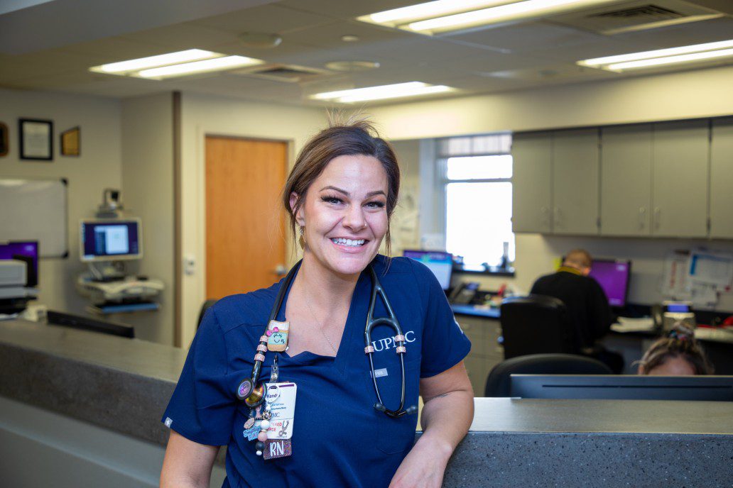Kendra wears blue scrubs at a hospital desk, looks at camera with a soft smile on her face.