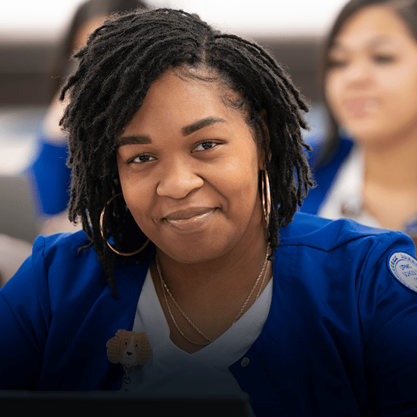 Nursing student smiles while in blue jacket and white scrubs.